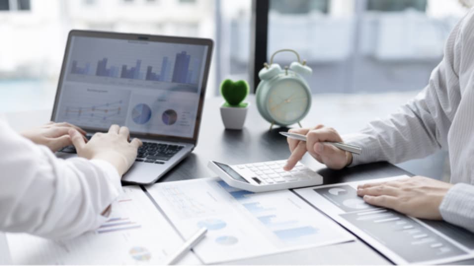 Laptop showing a bar chart next to hands writing notes with a calculator and small plant on a desk